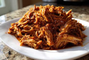 A close-up of tender, saucy crock pot pulled pork piled high on a white plate, glistening in the sunlight.