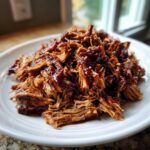 Close-up of tender, saucy crock pot pulled pork piled high on a white plate.