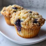 Two freshly baked blueberry muffins with a thick, sugary crumb topping sitting on a white plate.