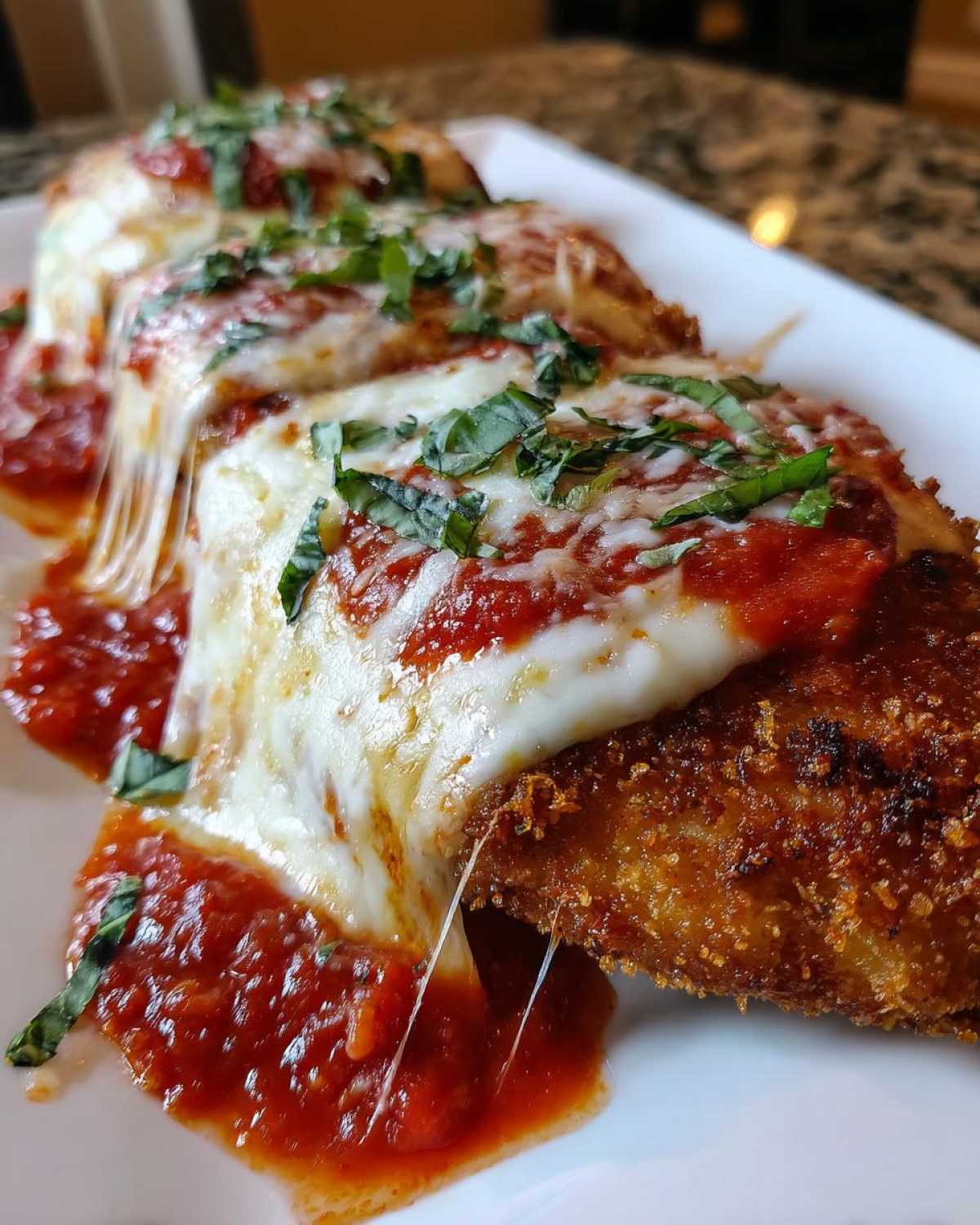 Close-up of three breaded chicken fillets topped with marinara sauce, melted mozzarella, and fresh basil, showing cheese pull.