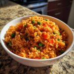 A white bowl filled with vibrant orange Spanish rice mixed with bright green peas and diced carrots, sitting on a granite countertop.