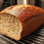 A freshly baked loaf of whole wheat bread, sliced to show the interior crumb, resting on a wire cooling rack.