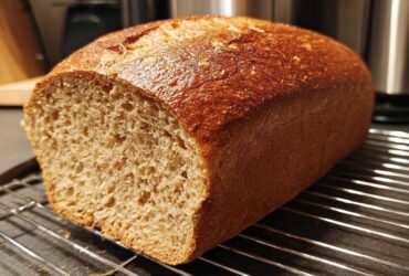 A freshly baked loaf of whole wheat bread, sliced to show the interior crumb, resting on a wire cooling rack.