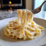 A fork lifts creamy fettuccine noodles coated in rich Olive Garden Alfredo sauce, topped with grated Parmesan cheese.
