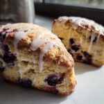 Two flaky blueberry scones drizzled generously with white vanilla glaze, sitting on a white marble surface.