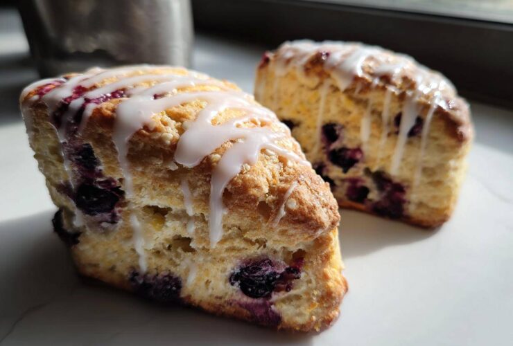 Two flaky blueberry scones drizzled generously with white vanilla glaze, sitting on a white marble surface.