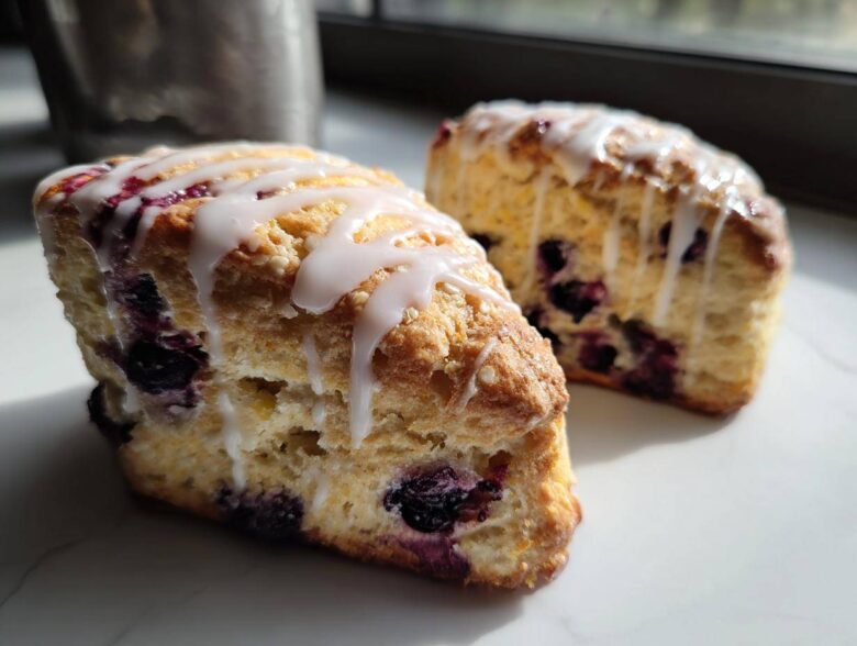 Two flaky blueberry scones drizzled generously with white vanilla glaze, sitting on a white marble surface.