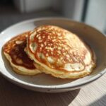 Two golden brown almond flour pancakes stacked on a speckled white plate, bathed in sunlight.