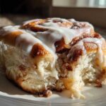 A close-up shot of a fluffy, freshly baked cinnamon roll covered in white icing, with a bite taken out.
