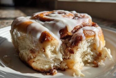 A close-up shot of a fluffy, freshly baked cinnamon roll covered in white icing, with a bite taken out.