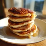 A tall stack of five fluffy sourdough pancakes dripping with golden syrup on a white plate.