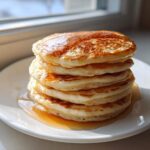 A tall stack of fluffy sourdough pancakes drizzled generously with golden syrup, sitting on a white plate.