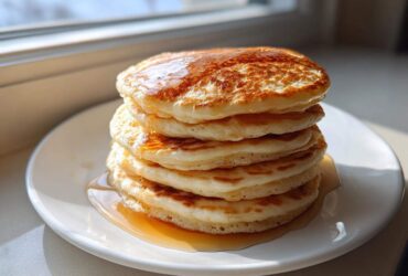 A tall stack of fluffy sourdough pancakes drizzled generously with golden syrup, sitting on a white plate.