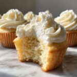 Close-up of a fluffy vanilla cupcakes with a bite taken out, showing the moist interior and rich white frosting.