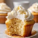 Close-up of a fluffy vanilla cupcake with a bite taken out, showing the moist crumb and rich white frosting.