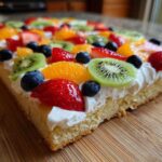 A close-up of a slice of fruit pizza sugar cookie topped with whipped cream, kiwi, strawberries, blueberries, and orange segments.