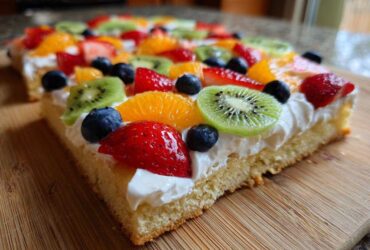 A close-up of a slice of fruit pizza sugar cookie topped with whipped cream, kiwi, strawberries, blueberries, and orange segments.