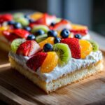 Close-up of a slice of fruit pizza sugar cookie topped with whipped cream, strawberries, kiwi, blueberries, and orange slices.