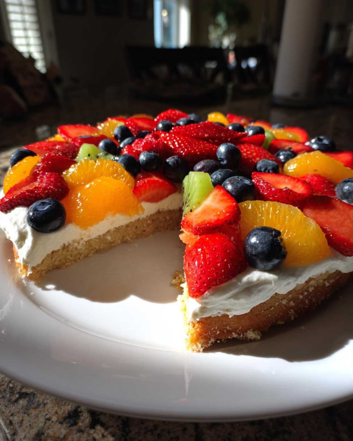 A vibrant fruit pizza sugar cookie with a slice cut out, topped with strawberries, blueberries, kiwi, and oranges.