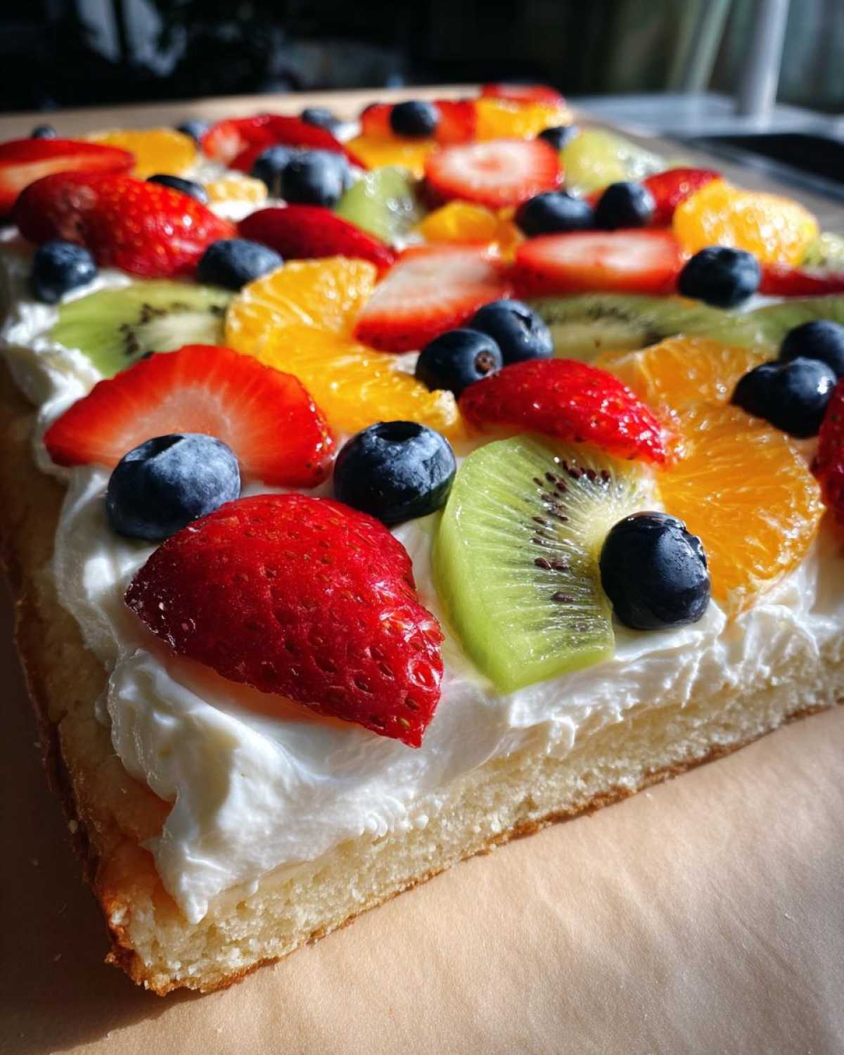 A close-up view of a slice of fruit pizza sugar cookie topped with white frosting, strawberries, blueberries, kiwi, and orange slices.