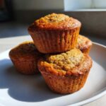 Three golden-brown banana protein muffins stacked on a white plate, highlighted by strong sunlight.