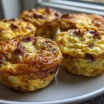 Close-up of four golden brown sausage Egg Muffin Cups resting on a light-colored plate near a window.