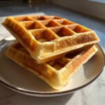 Two golden brown, fluffy high protein waffles stacked on a white plate near a window.