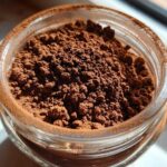 Close-up overhead view of rich, dark brown homemade hot cocoa mix powder filling a small glass jar.