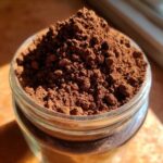 Close-up of rich, dark brown homemade hot cocoa mix powder mounded high in a small glass jar.