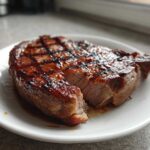 Close-up of a perfectly grilled and juicy pork steak resting on a white plate, showing clear grill marks.