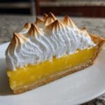 A close-up of a slice of lemon meringue pie showing bright yellow filling and tall, toasted meringue peaks.