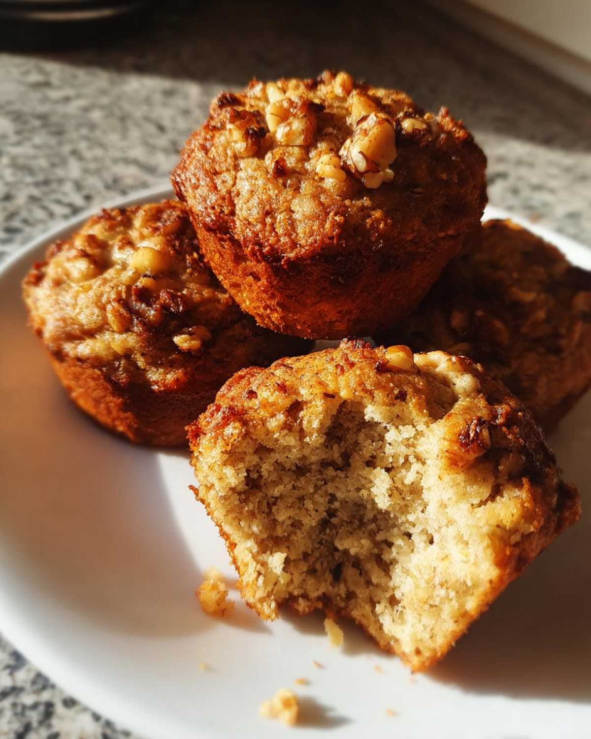 A stack of golden brown banana nut muffins, one of which has a bite taken out showing the moist interior and walnut topping.
