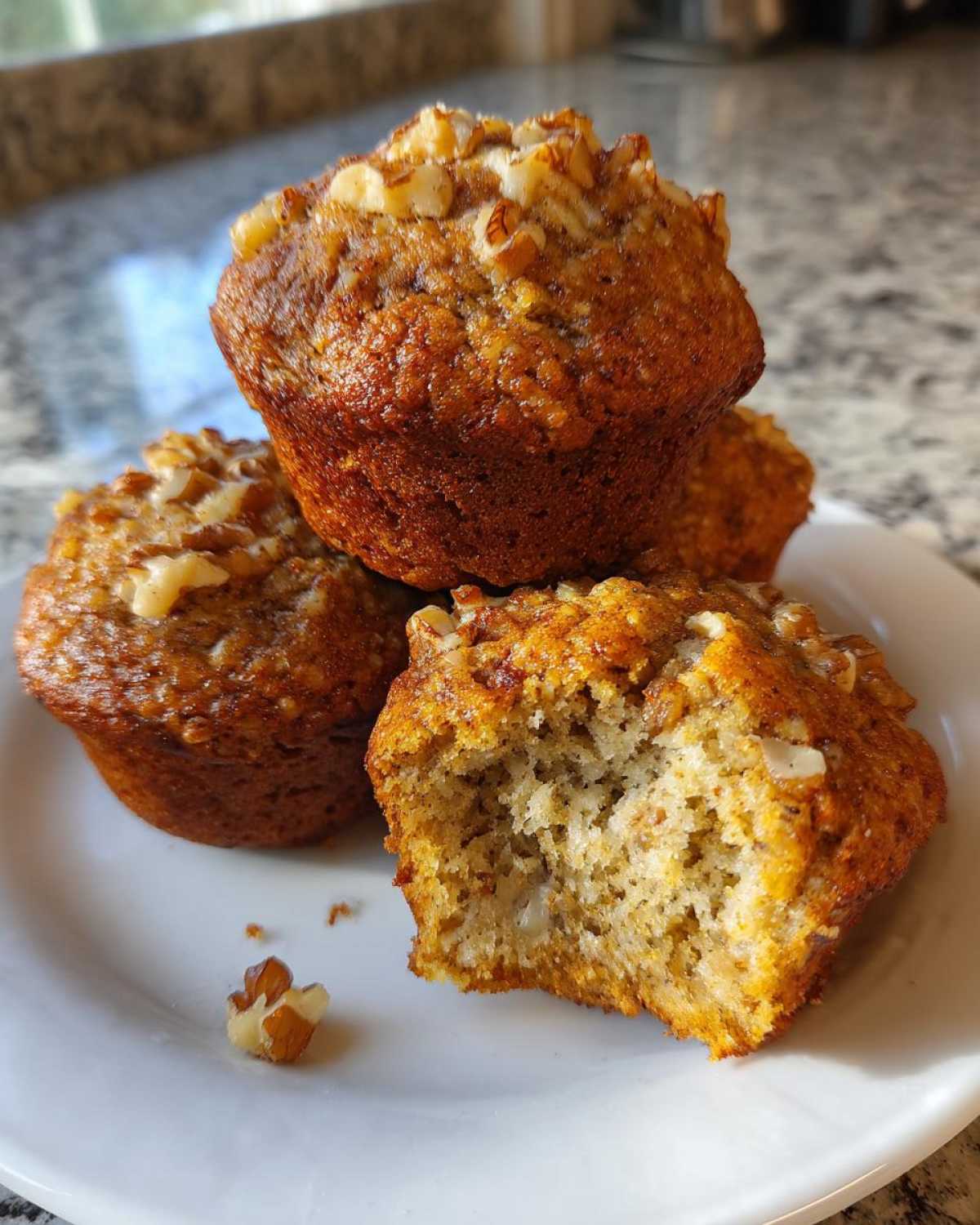 A stack of moist banana nut muffins topped with walnuts, with one muffin having a bite taken out showing the fluffy interior.