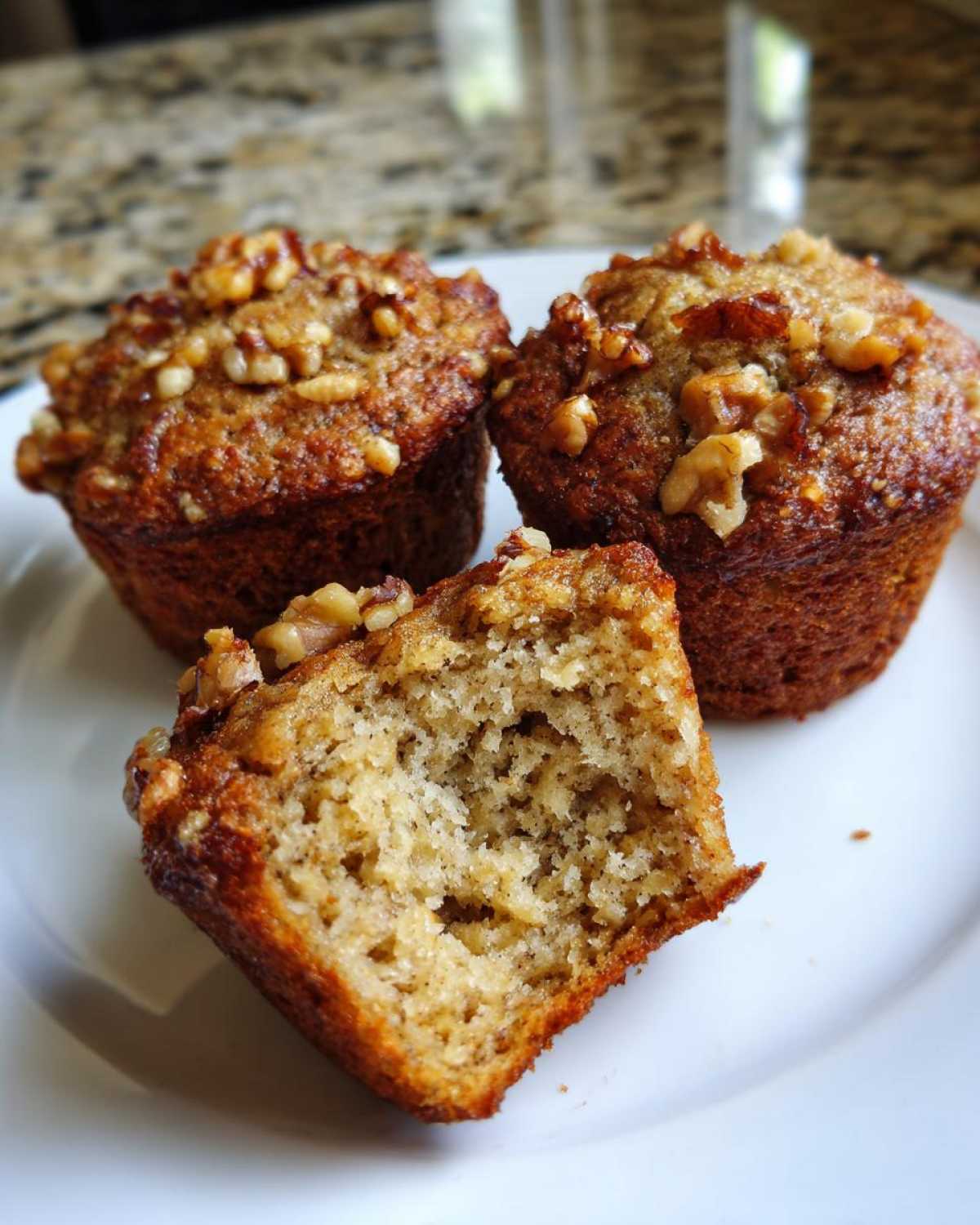 Three freshly baked banana nut muffins on a white plate, one cut open to show the moist interior.