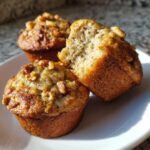 Three freshly baked banana nut muffins on a white plate, one is broken open showing the moist interior texture.