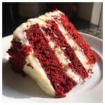 A close-up, appetizing slice of moist red velvet cake showing three rich red layers separated by thick white cream cheese frosting.
