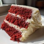 Close-up of a moist slice of red velvet cake featuring three layers of deep red cake separated by thick white cream cheese frosting.