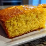 Close-up of a moist, golden slice of sourdough cornbread with a shiny glaze on top, served on a white plate.