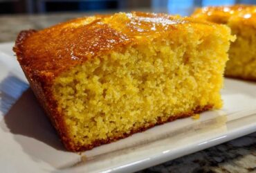 Close-up of a moist, golden slice of sourdough cornbread with a shiny glaze on top, served on a white plate.