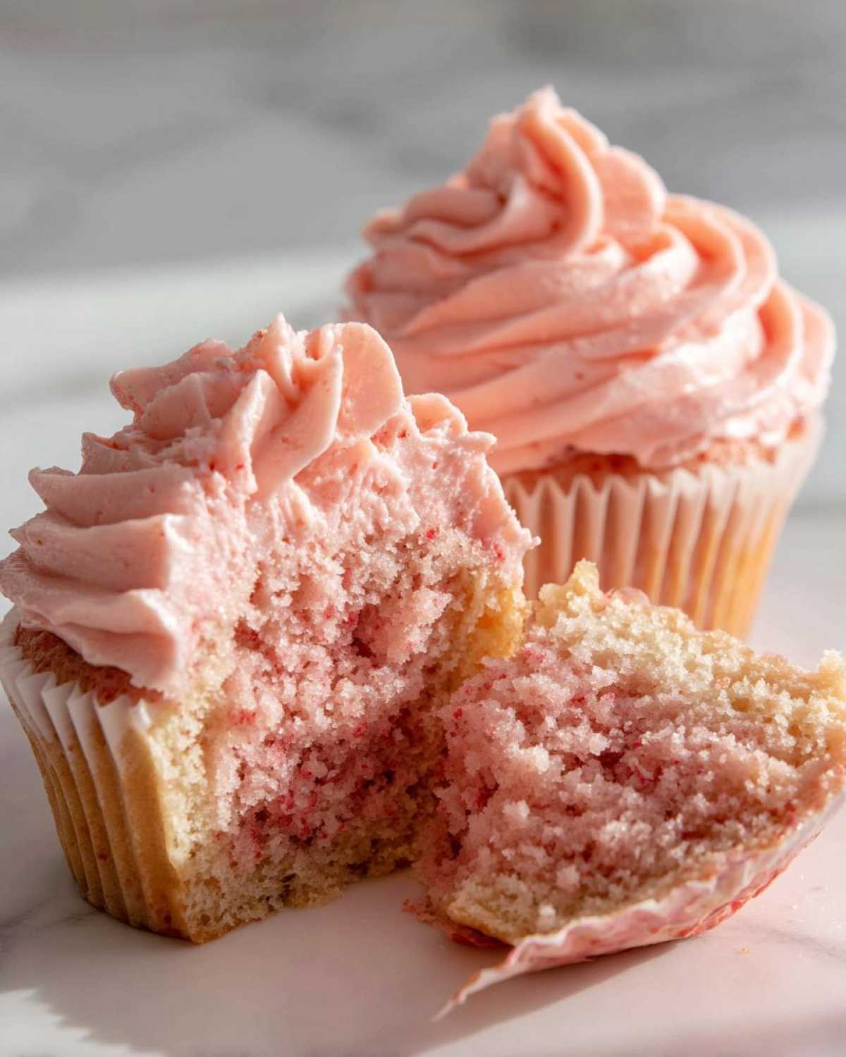 Close-up of a strawberry cupcake cut in half showing the moist, pink-flecked crumb and swirled pink frosting.
