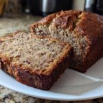 Close-up of a moist, dark-crusted sugar free banana bread loaf with one slice cut and resting beside it on a white plate.