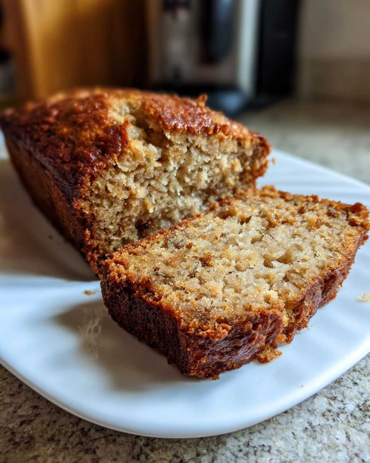 A loaf of moist sugar free banana bread with one thick slice cut and resting against the main loaf on a white plate.