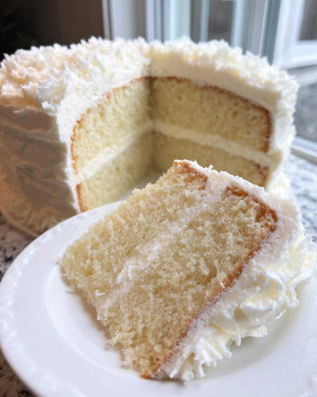 A close-up of a slice of moist vanilla cake recipe, layered with white frosting, next to the rest of the cake.