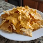 A white plate piled high with tortilla chips generously covered in smooth, bright orange nacho cheese sauce.