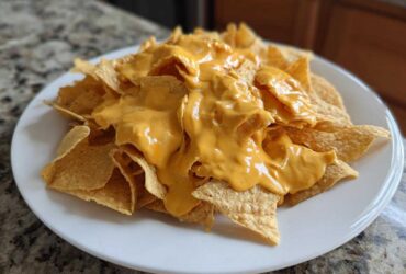 A white plate piled high with tortilla chips generously covered in smooth, bright orange nacho cheese sauce.
