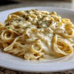 Close-up of fettuccine pasta generously coated in rich Olive Garden Alfredo Sauce and sprinkled with dried herbs.