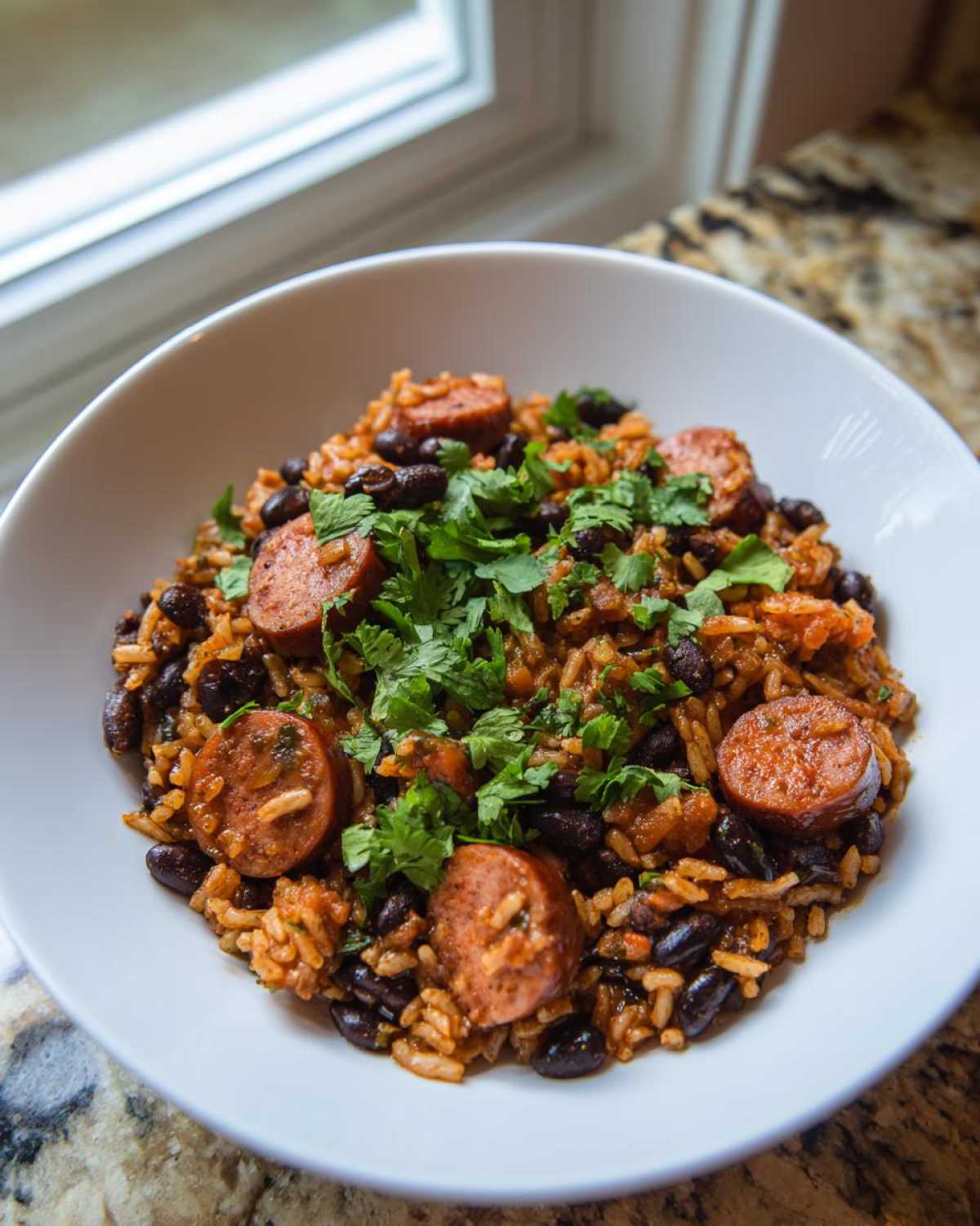A white bowl filled with flavorful black beans and rice mixed with sliced sausage and topped with fresh cilantro.