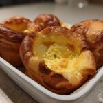 A close-up of several golden brown, perfectly risen Yorkshire pudding baked in a white rectangular dish.