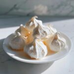 A close-up of several perfectly piped, crisp white meringue cookies piled on a small white plate.