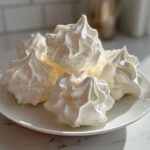 A close-up of several perfectly piped, white meringue cookies stacked on a small white plate.